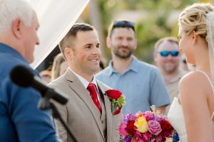 Groom and bride exchanging vows at outdoor wedding ceremony, with family and friends, beautiful flowers, and wedding officiant present.