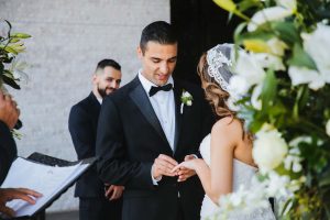 Elegant wedding ceremony with groom and bride exchanging vows, surrounded by floral decor and officiant, captured by EMweddings.