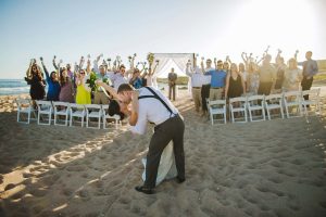 Beach wedding ceremony with newlyweds kissing and guests celebrating on the sand at sunset.
