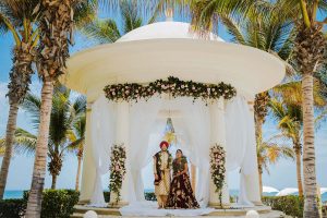 Elegant beachside wedding ceremony with floral decorations, under a white gazebo surrounded by palm trees.