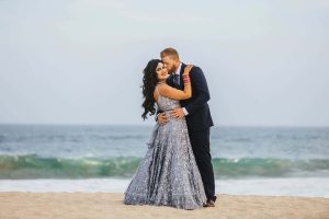 Elegant couple in wedding attire embrace on the beach with ocean waves in the background, celebrating love and marriage.