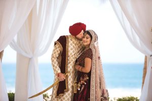Elegant Indian wedding couple sharing a romantic moment under a white draped archway near the beach.